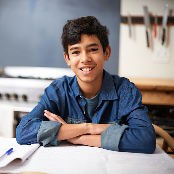 A young boy sits at a table with his arms crossed, smiling